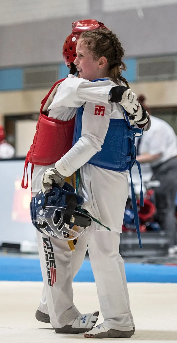 Zwei Taekwondo-Athleten, einer mit rotem Kopfschutz und einer mit blauem, umarmen sich nach einem Kampf. Beide tragen Schutzkleidung und Handschuhe.