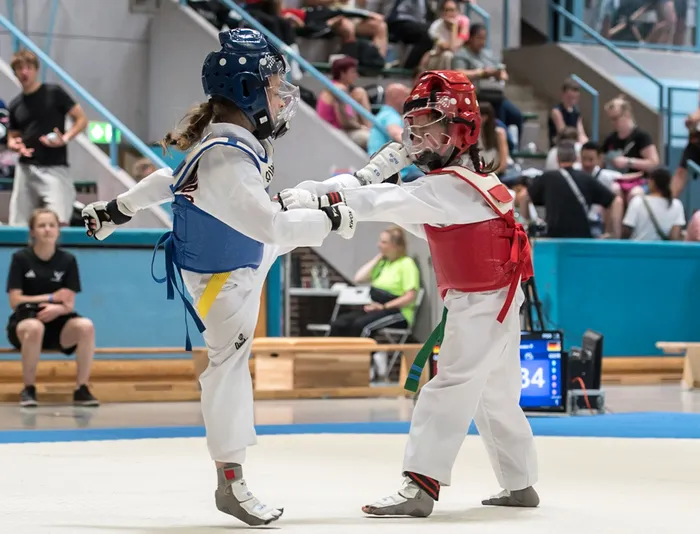 Zwei Taekwondo-Athletinnen in voller Schutzausrüstung kämpfen in einer Halle. Die Athletin links trägt Blau, die rechts Rot. Im Hintergrund sind Zuschauer und eine Anzeigetafel zu sehen.