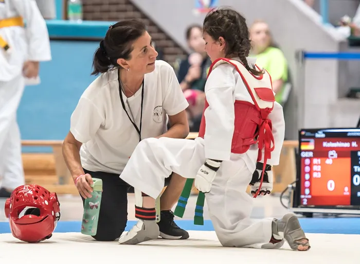 Eine Trainerin kniet neben einer jungen Taekwondo-Athletin und justiert ihre Fußschützer. Im Hintergrund sind andere Sportler und eine Anzeigetafel zu sehen.