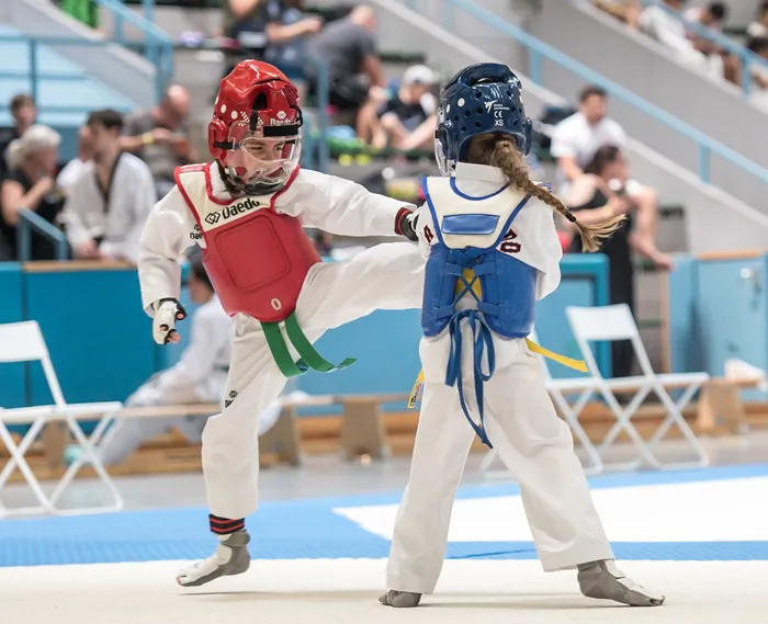 Zwei junge Taekwondo-Kämpferinnen in voller Montur treten gegeneinander an. Die Kämpferin in Rot führt einen Tritt aus, während ihre Gegnerin in Blau sich verteidigt. Im Hintergrund sind Zuschauer und die Sporthalle zu sehen.