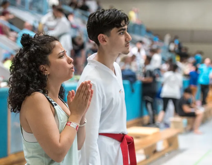 Zwei junge Menschen stehen nebeneinander und schauen konzentriert zu. Die Frau betet mit gefalteten Händen, während der Mann ein weißes Taekwondo-Uniform und einen roten Gürtel trägt. Im Hintergrund sind weitere Personen und eine Sporthalle zu sehen.