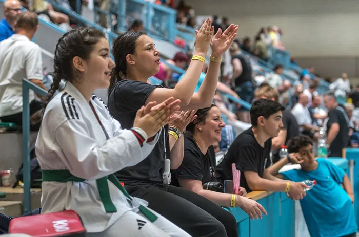 Zuschauer klatschen und jubeln bei einem Taekwondo-Turnier in einer Sporthalle. Im Vordergrund zwei junge Frauen, eine in Kampfmontur mit grünem Gürtel.