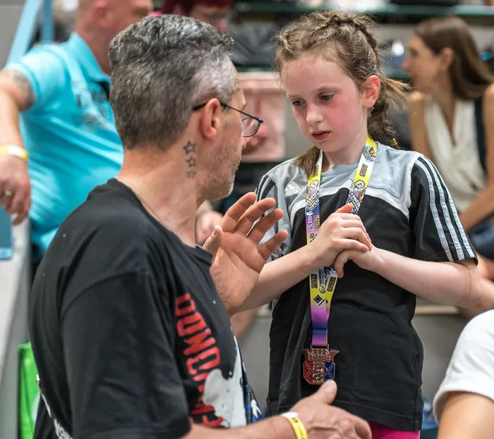 Ein Mann mit Tätowierungen und ein rotes T-Shirt spricht mit einem jungen Mädchen, das mehrere Medaillen um den Hals trägt. Im Hintergrund sind weitere Personen und eine Sporthalle zu sehen.