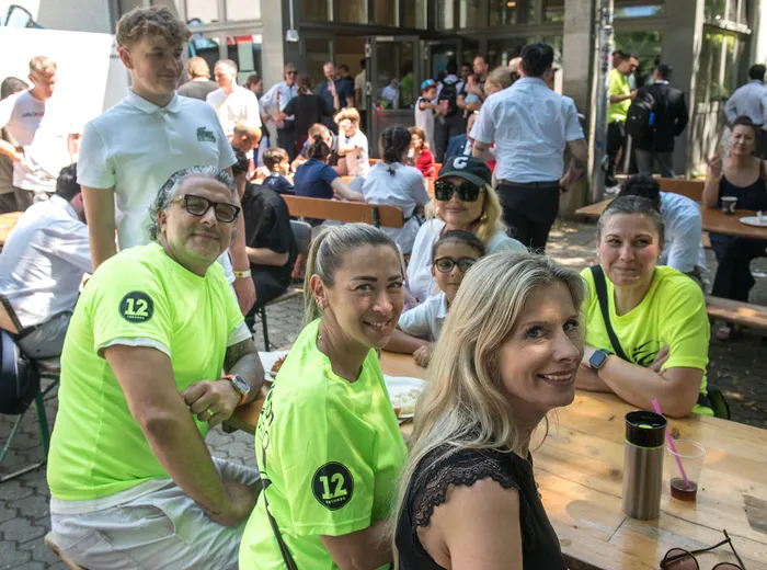 Eine Gruppe von Menschen sitzt an Holztischen im Freien, einige tragen grüne Trikots mit der Nummer 12. Im Hintergrund sind weitere Personen und ein Gebäude zu sehen.
