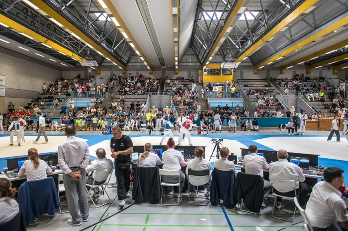 Zwei Taekwondo-Kämpfer in weißen Anzügen stehen auf einer blauen Matte in einer großen Sporthalle vor einer Zuschauertribüne. Vor den Zuschauern sitzen Kampfrichter an Tischen mit Bildschirmen.