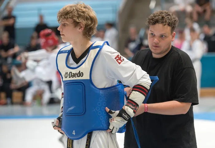 Ein junger Taekwondo-Athlet mit blonder Frisur und Schutzausrüstung wird von einem Betreuer mit dunklen Haaren und schwarzem T-Shirt am Spielfeldrand unterstützt. Im Hintergrund sind weitere Sportler und Zuschauer zu sehen.