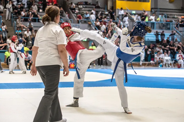 Zwei Taekwondo-Athleten kämpfen in einem Turnier, während eine Schiedsrichterin das Geschehen beobachtet. Die Sportlerin in Blau führt einen Tritt aus.
