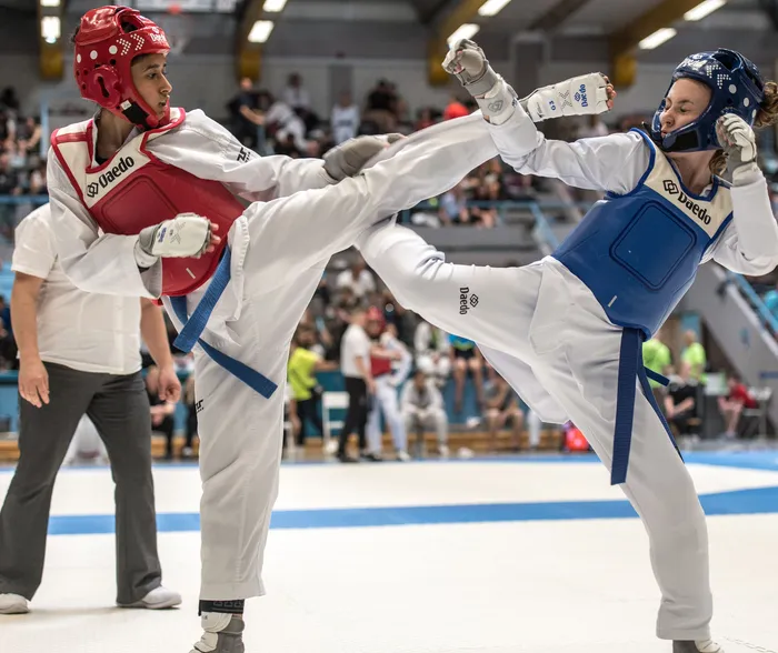Zwei Taekwondo-Athleten, eine in roter Schutzausrüstung und eine in blauer, kämpfen in einem Wettkampf. Die Athletin in Blau führt einen Tritt aus, während ihre Gegnerin in roter Ausrüstung blockt.