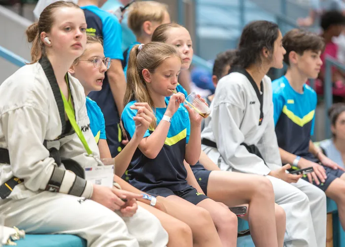 Junge Sportlerinnen und Sportler sitzen auf einer Bank und trinken oder warten auf ihren Einsatz beim Taekwondo-Turnier. Einige tragen Schutzausrüstung und Headsets.