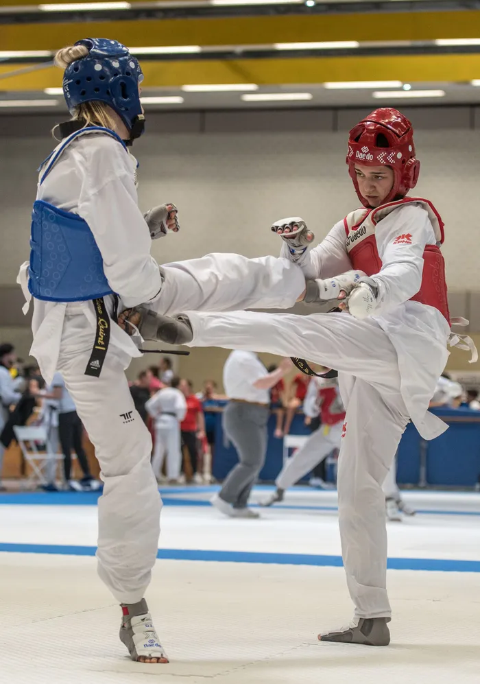 Zwei Taekwondo-Athleten in voller Schutzausrüstung kämpfen in einer Halle. Der Athlet rechts führt einen Tritt aus, während die linke Athletin blockt.