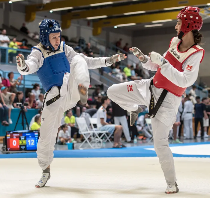 Zwei Taekwondo-Athletinnen kämpfen in einem Wettkampf. Die Sportlerin links trägt einen blauen Schutzanzug, die rechts einen roten. Im Hintergrund sind Zuschauer und eine Anzeigetafel zu sehen.