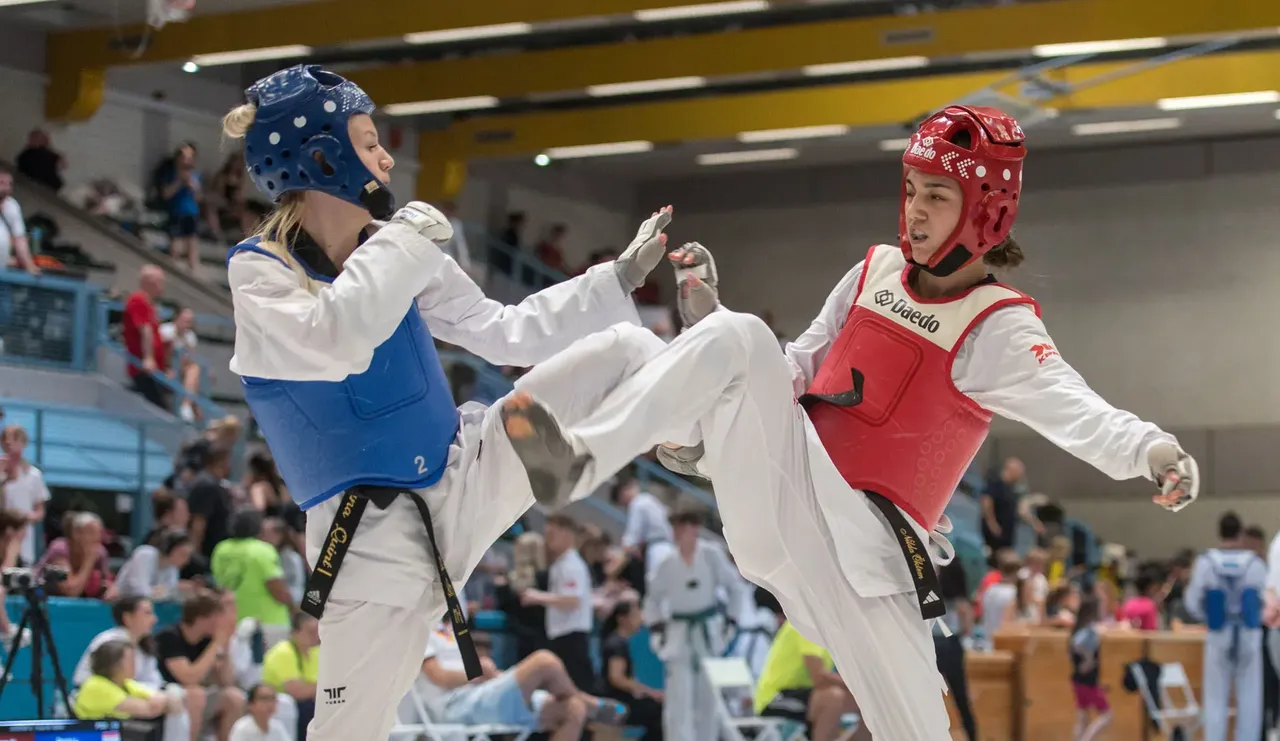 Zwei Taekwondo-Athleten in voller Schutzausrüstung kämpfen in einem Wettkampf. Die Sportlerin in Rot führt einen Tritt aus, während ihre Gegnerin in Blau blockt.
