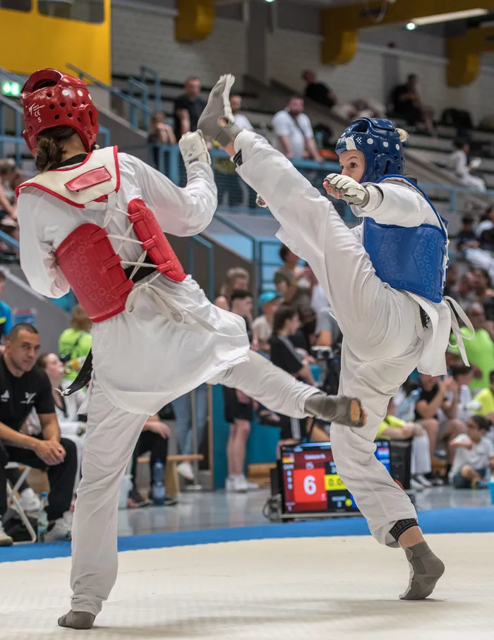 Zwei Taekwondo-Athletinnen in voller Schutzausrüstung treten in einem Wettkampf gegeneinander an. Die Athletin rechts führt einen Tritt aus, während ihre Gegnerin ausweicht. Im Hintergrund sind Zuschauer und eine Anzeigetafel zu sehen.