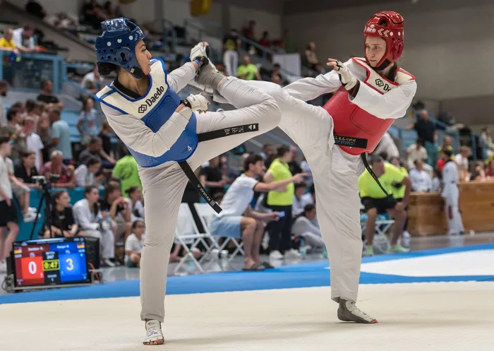 Zwei Taekwondo-Athletinnen in voller Montur treten in einem Wettkampf gegeneinander an. Die Athletin links trägt einen blauen Anzug, die rechts einen roten. Im Hintergrund sind Zuschauer und eine Anzeigetafel zu sehen.