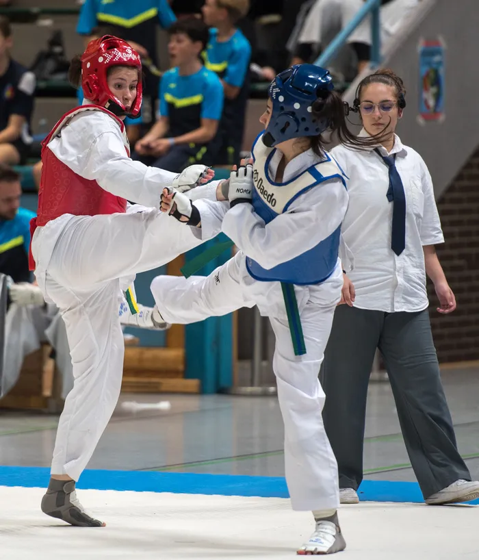 Zwei Taekwondo-Athletinnen in voller Montur kämpfen in einer Halle. Die Athletin links führt einen Tritt aus, während die Athletin rechts blockt. Im Hintergrund steht eine Schiedsrichterin.