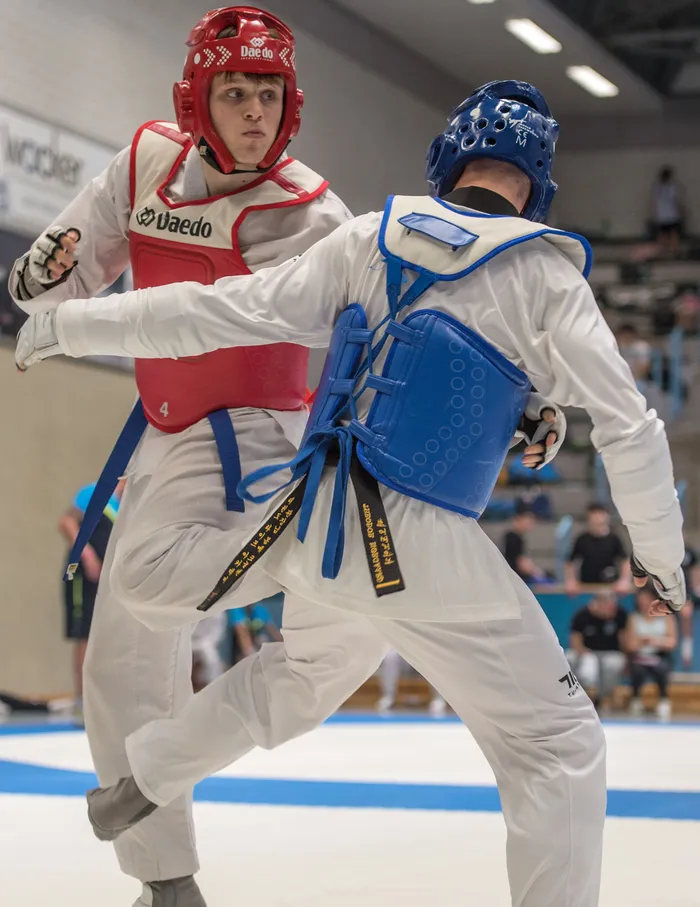 Zwei Taekwondo-Athleten in voller Schutzausrüstung kämpfen in einer Halle. Der Athlet links trägt einen roten Helm und ein rotes Brustschutz, der Athlet rechts einen blauen Helm und einen blauen Brustschutz. Im Hintergrund sind Zuschauer zu sehen.