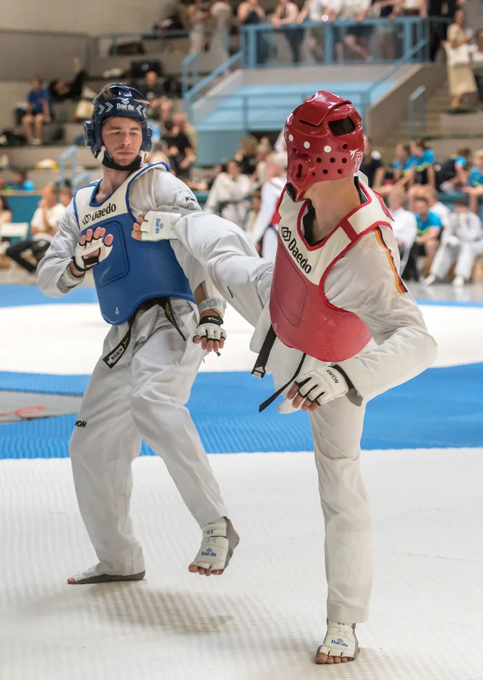 Zwei Taekwondo-Athleten in voller Schutzausrüstung kämpfen in einer Halle vor Zuschauern. Der Athlet rechts trägt einen roten Helm und eine Schutzausrüstung mit den Farben des deutschen Bundesadlers.