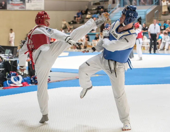 Zwei Taekwondo-Athleten in voller Schutzausrüstung kämpfen in einem Wettkampf. Der Athlet im blauen Anzug führt einen Tritt aus, während sein Gegner im roten Anzug blockt.