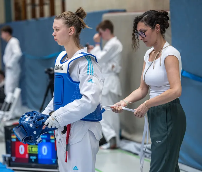 Eine Taekwondo-Athletin in blauer Schutzausrüstung steht neben einer Trainerin, die ihr die Ausrüstung anpasst. Im Hintergrund sind weitere Sportler und eine Anzeigetafel zu sehen.