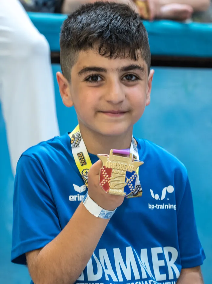 Ein lächelnder Junge hält eine goldene Medaille mit einer blauen Schleife in der Hand. Er trägt ein blaues Sporttrikot mit Werbeaufdruck und eine Medaille um den Hals.