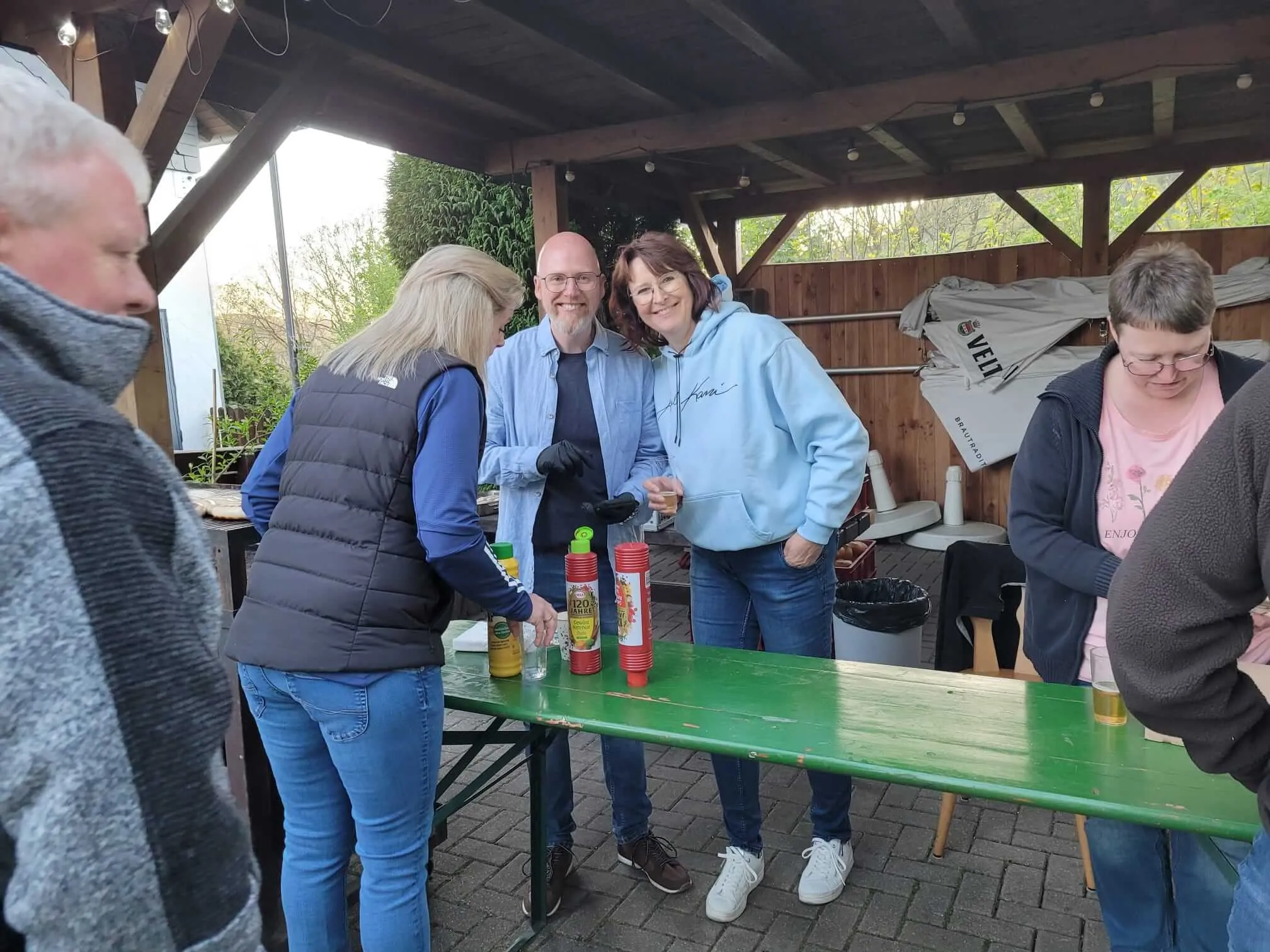 Drei Personen stehen an einem Tisch mit verschiedenen Soßen und Getränken im Freien, im Hintergrund weitere Menschen und ein überdachter Bereich.