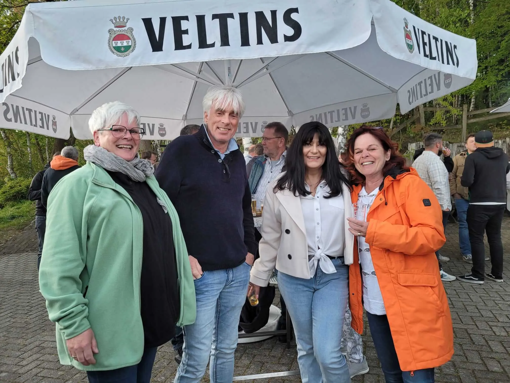 Vier lächelnde Menschen stehen vor Schirmständern mit dem Velttins-Logo. Im Hintergrund sind weitere Menschen und Bäume zu sehen.
