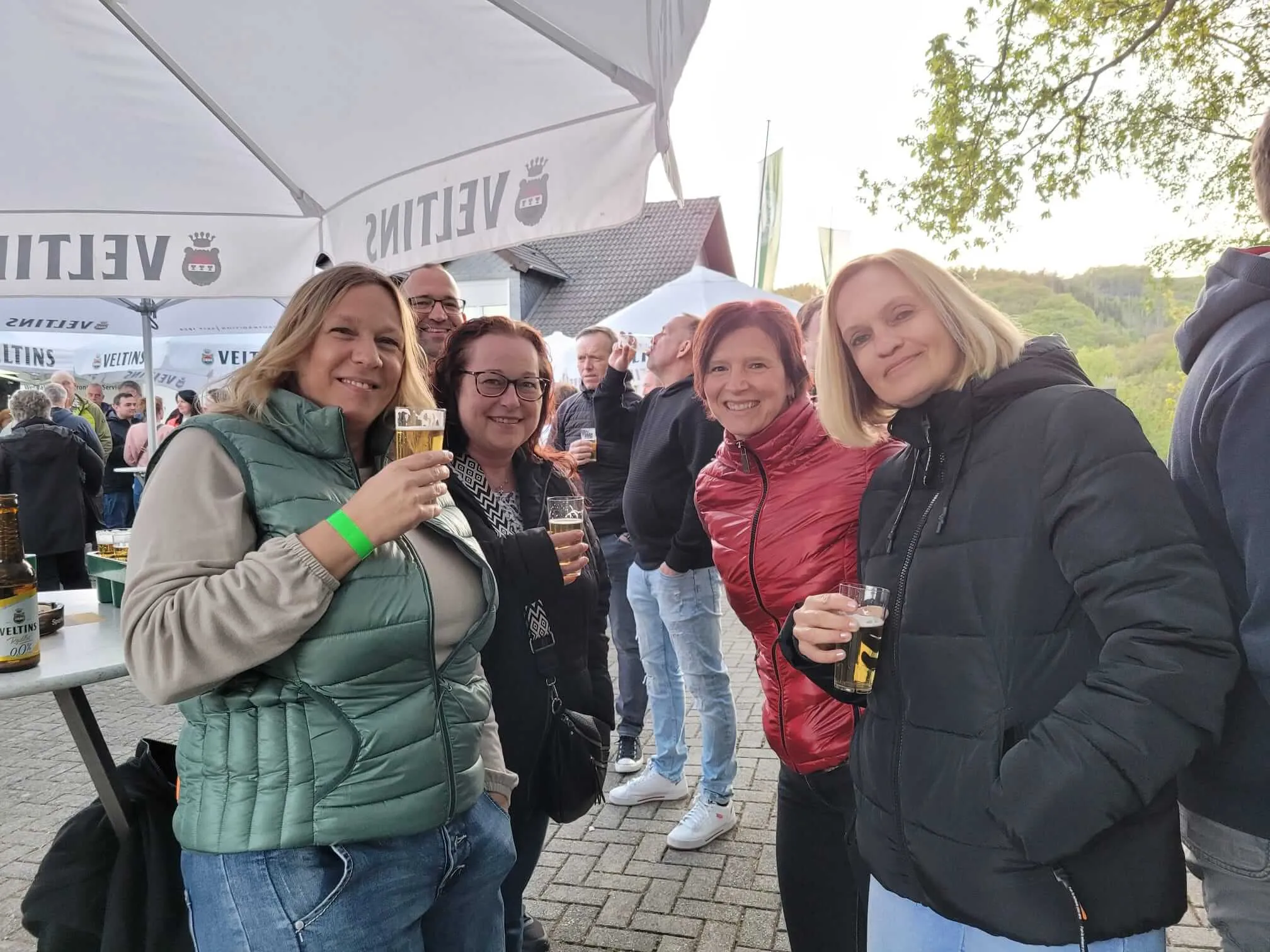Mehrere lächelnde Menschen stehen eng zusammen und halten Biergläser in der Hand, im Hintergrund ein Pavillon mit dem Schriftzug „Veitins“.
