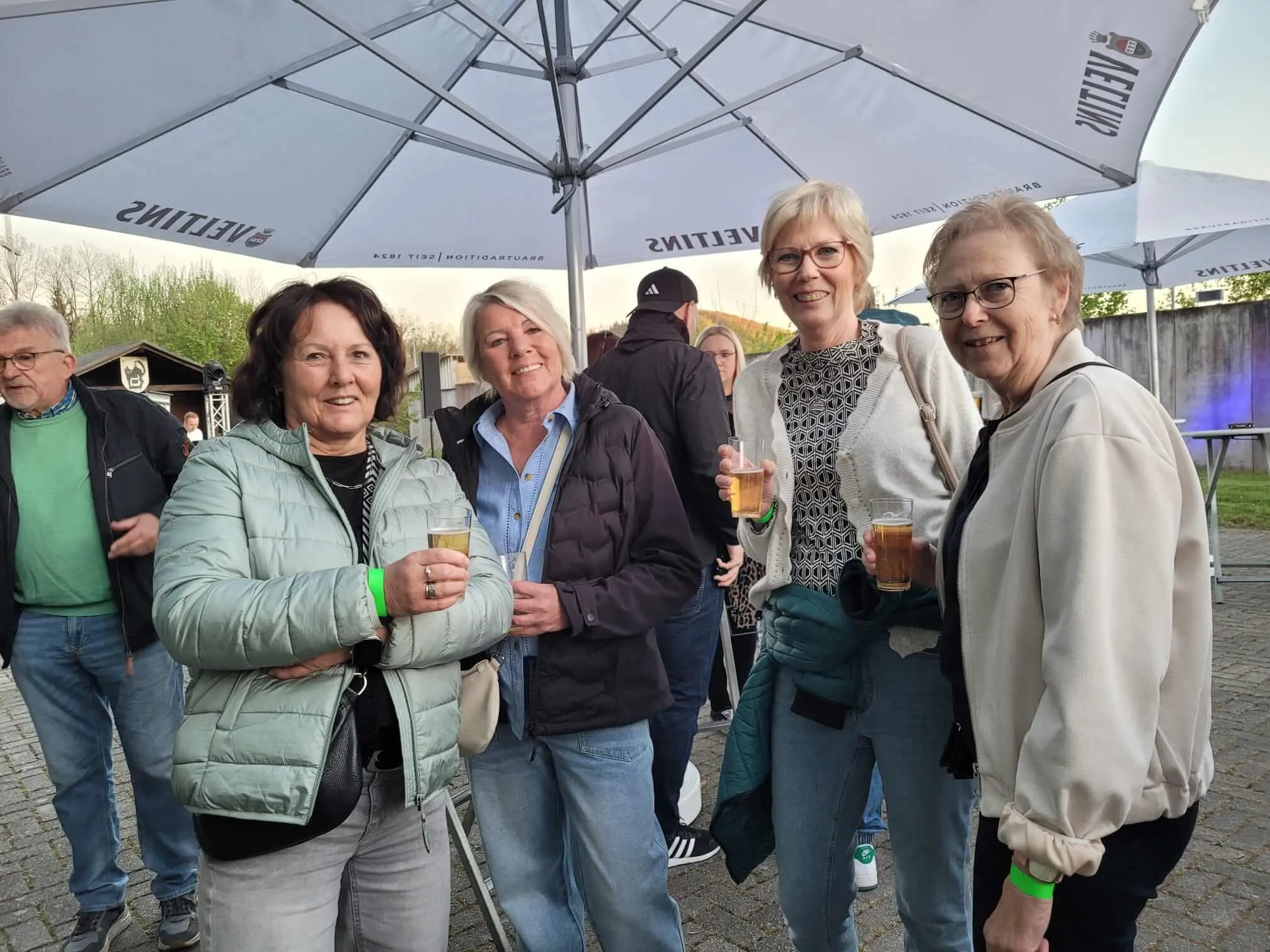 Eine Gruppe von Menschen steht unter einem großen Sonnenschirm und hält Biergläser in der Hand. Im Hintergrund sind weitere Personen und Tische zu sehen.