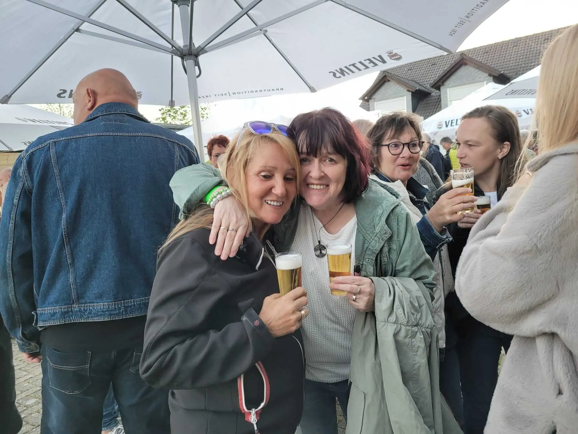 Mehrere Menschen stehen eng zusammen und lächeln in die Kamera, einige halten Biergläser. Im Hintergrund sind Zelte und weitere Personen zu sehen.