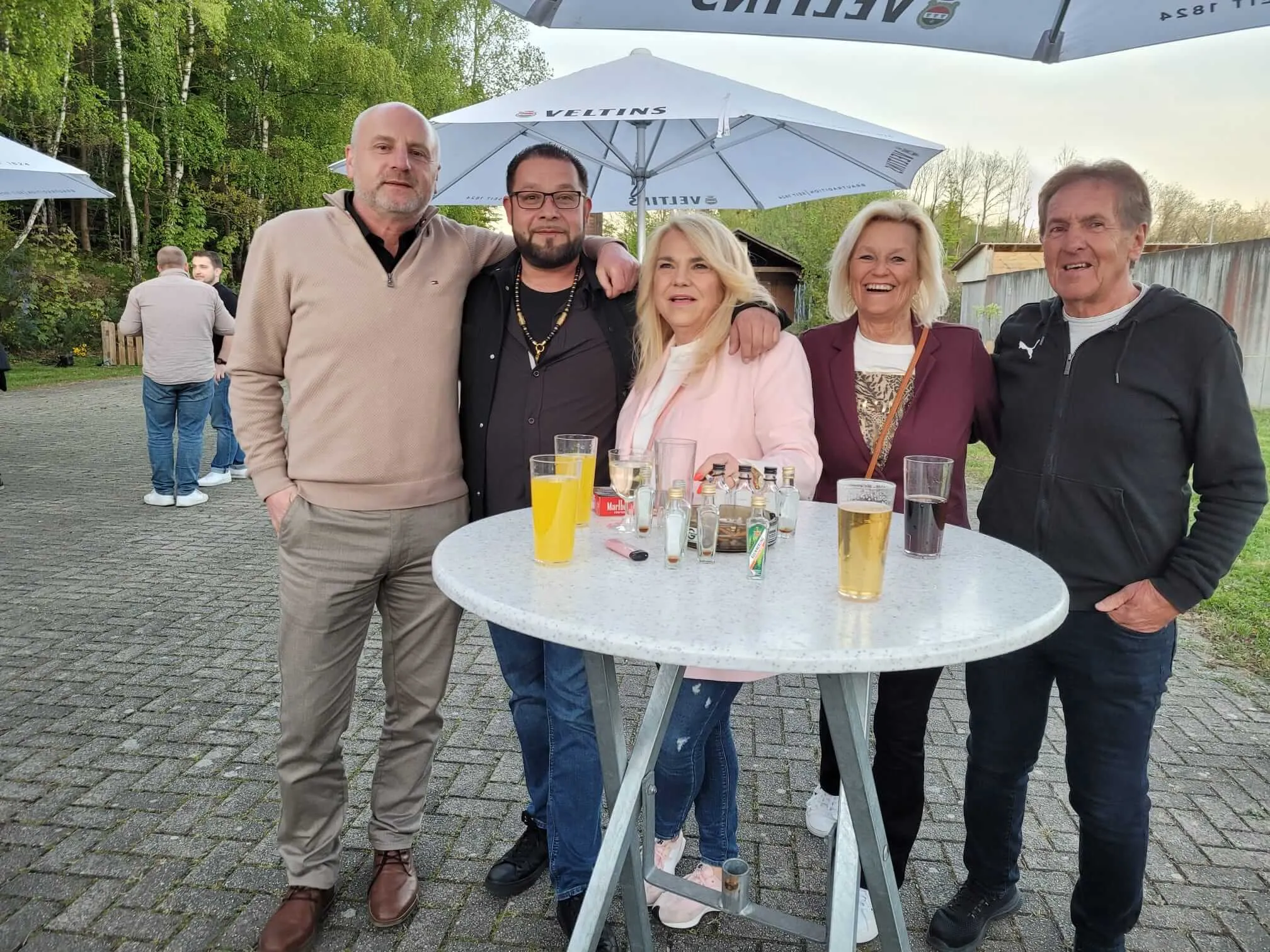 Fünf Personen stehen um einen runden Tisch im Freien, Getränke vor sich. Im Hintergrund weitere Menschen und Sonnenschirme.