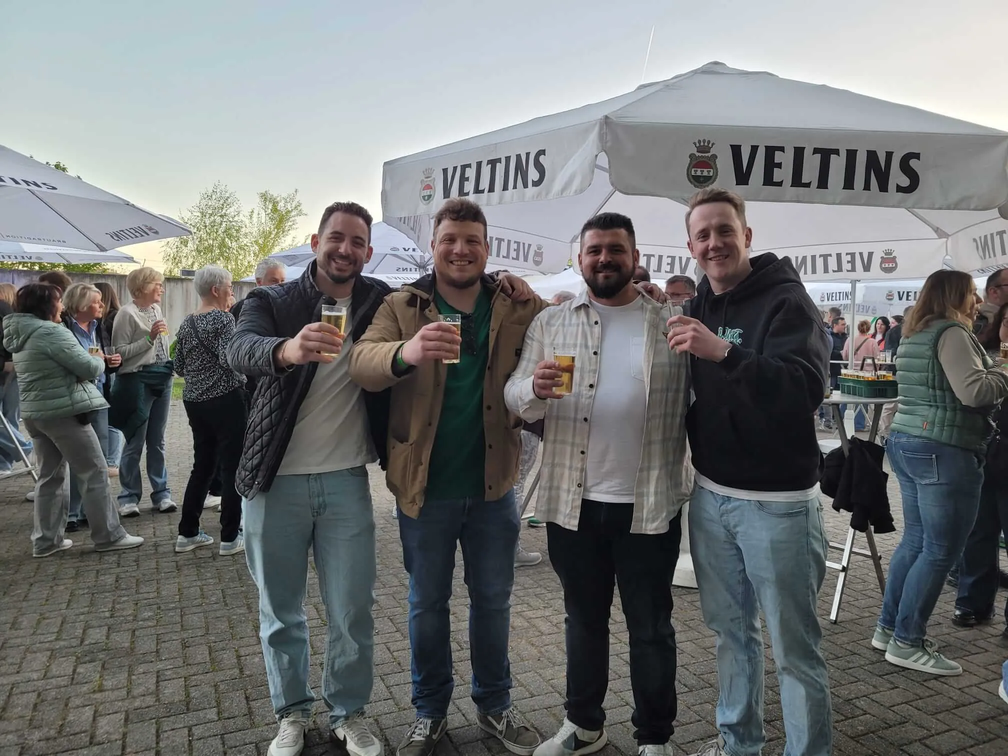 Drei Männer heben gefüllte Biergläser vor Zelten mit dem Velttins-Logo beim Tanz in den Mai des Schützenvereins Eiringhausen.