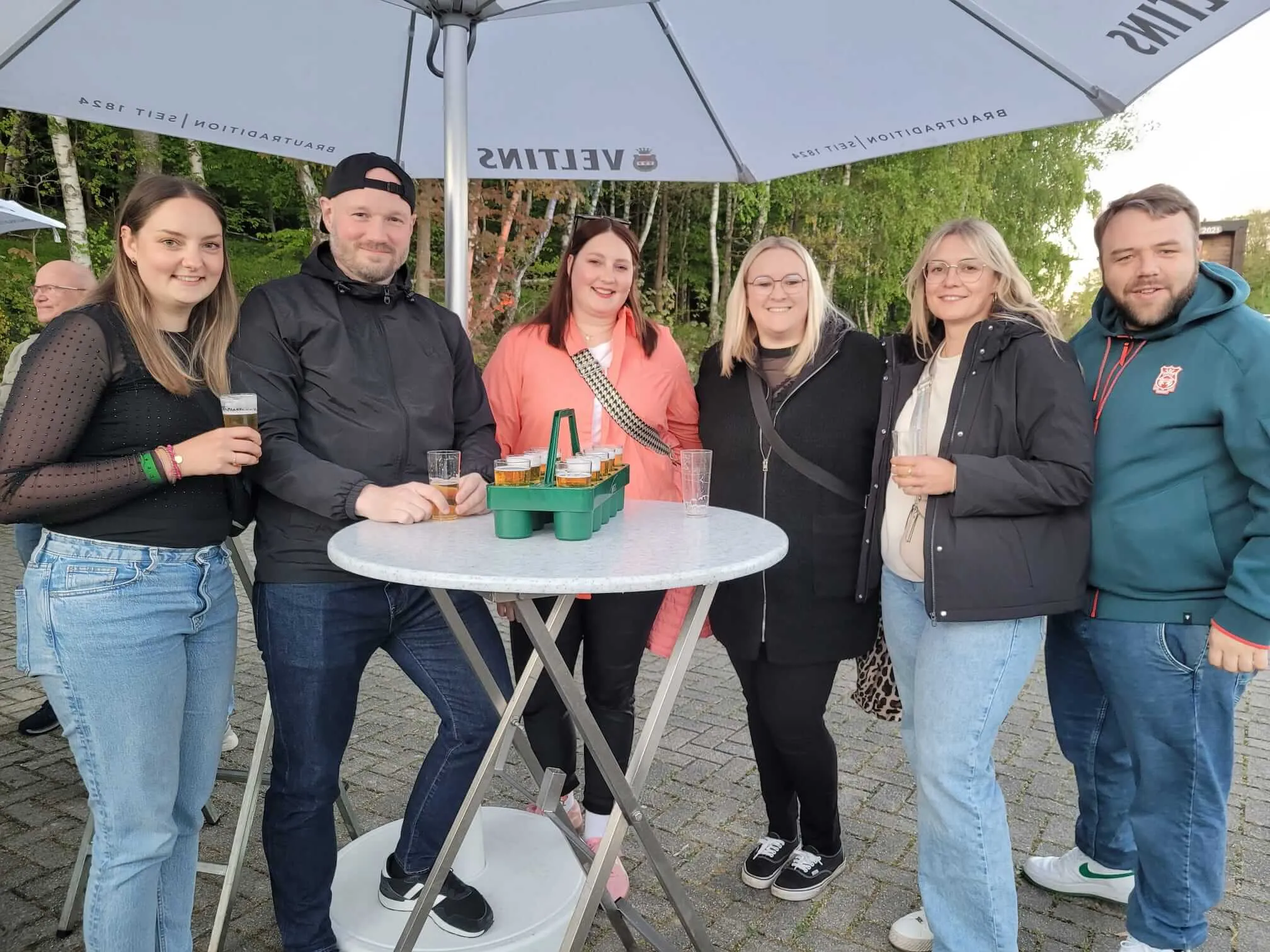 Sechs Personen stehen um einen runden Tisch unter Sonnenschirmen, einige halten Biergläser. Im Hintergrund sind weitere Menschen zu sehen.