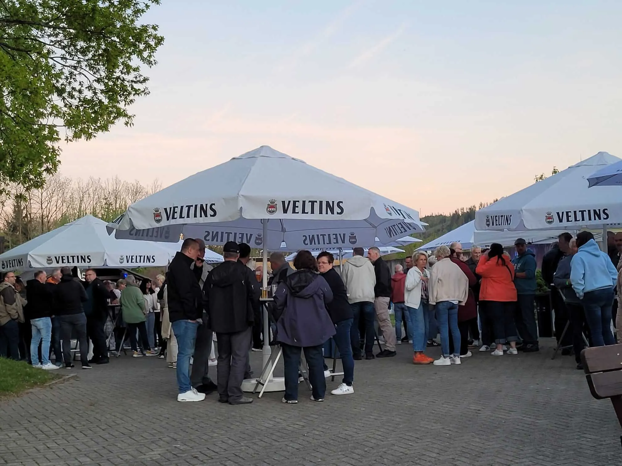 Menschen stehen und unterhalten sich vor mehreren weißen Zelten mit dem Aufdruck „VELTINS“ im Freien.