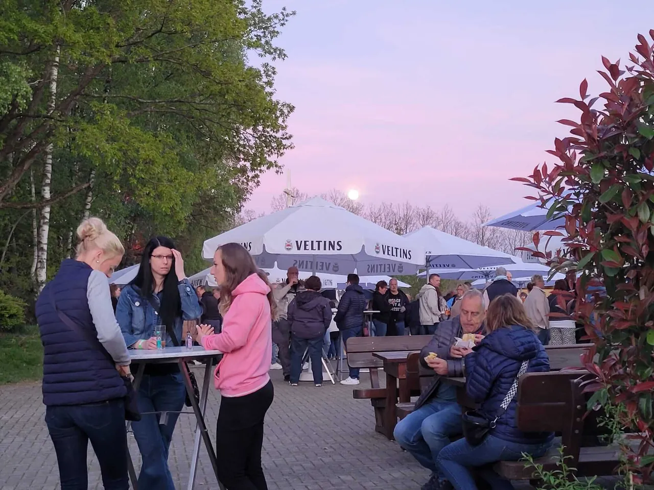Menschen stehen und sitzen im Freien vor mehreren Zelten mit dem Schriftzug „VELTINS“ bei einem Abendevent mit Abendhimmel im Hintergrund.