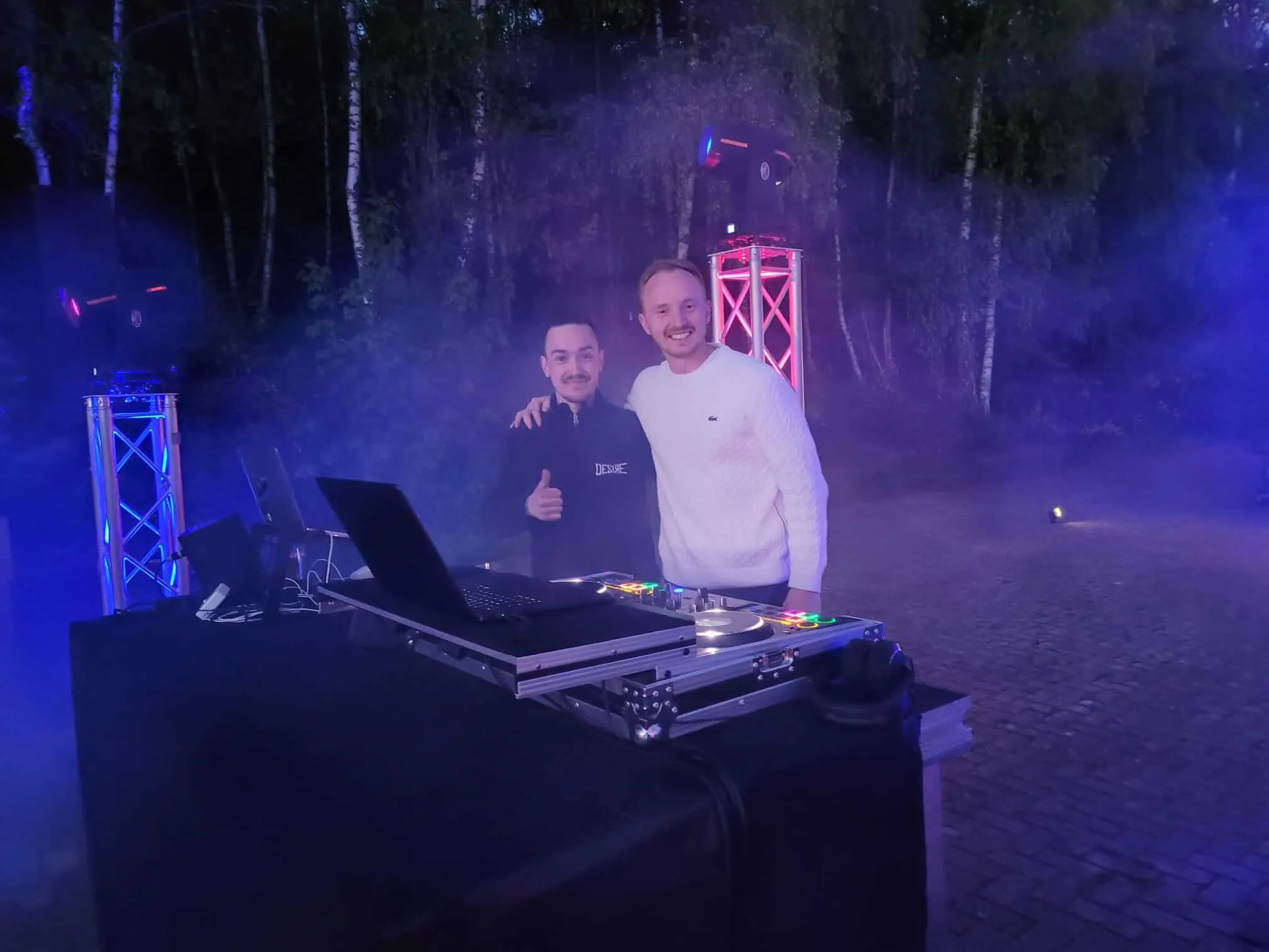 Zwei Männer stehen vor einer DJ-Anlage im Freien, umgeben von Bäumen. Der Mann links zeigt Daumen hoch, der andere lächelt in die Kamera.