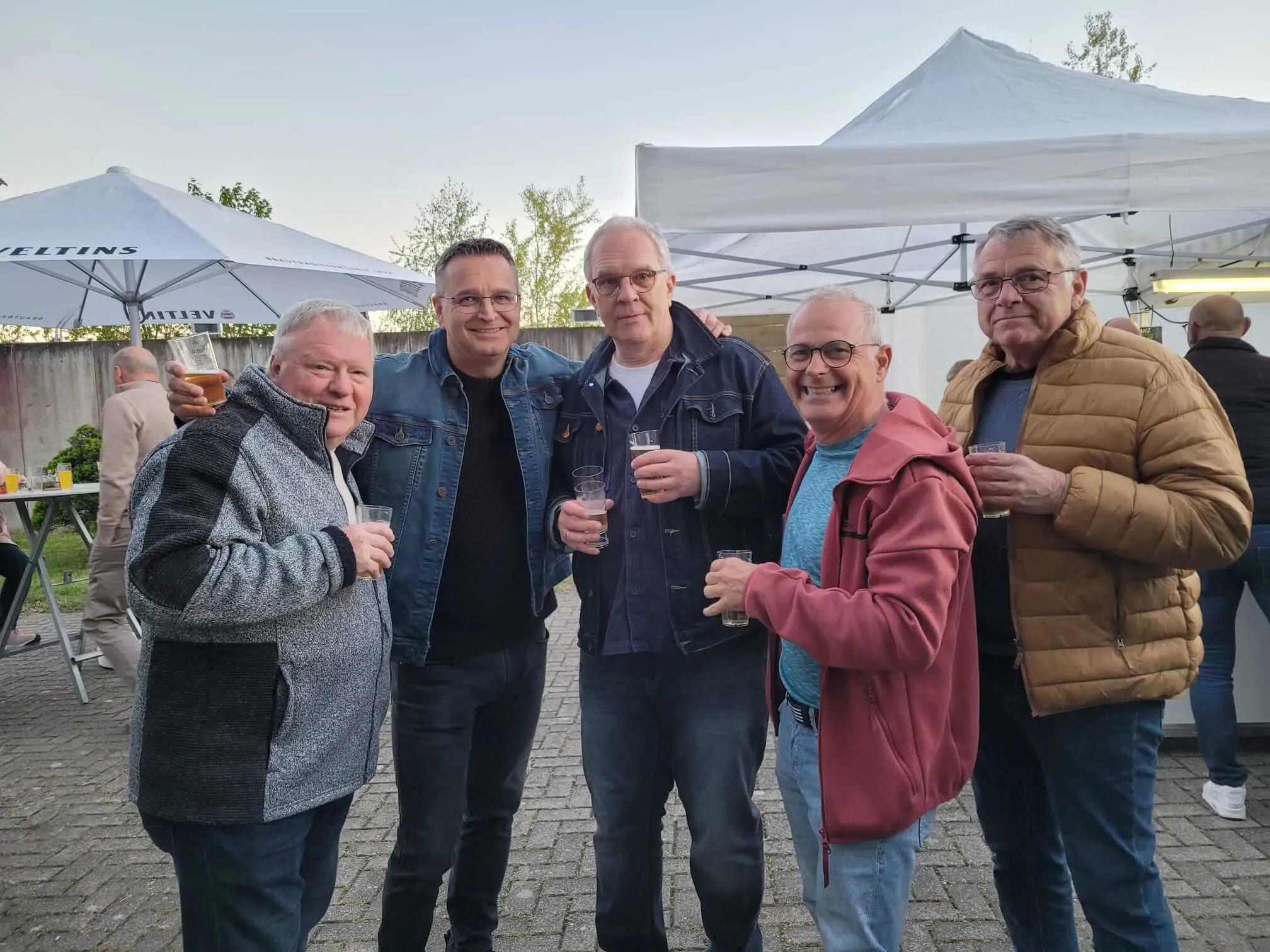Fünf Männer stehen lächelnd eng zusammen und halten Biergläser in der Hand. Im Hintergrund ein Zelt und weitere Personen.