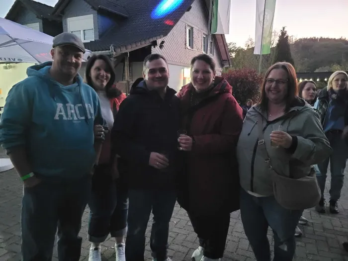 Fünf lächelnde Menschen stehen eng zusammen vor einem Gebäude mit festlicher Beleuchtung. Im Hintergrund weitere Personen und ein Zelt.