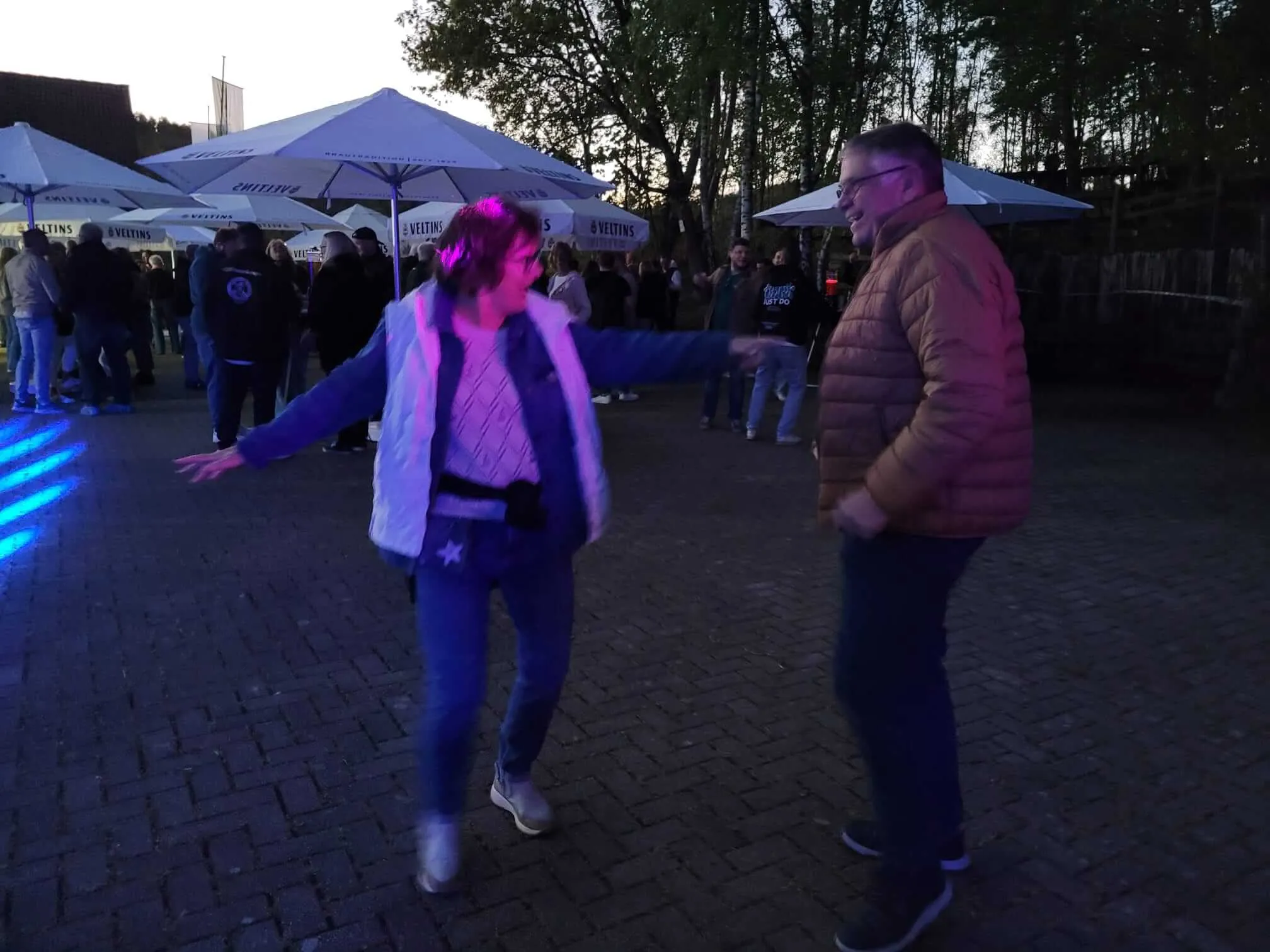 Zwei Menschen tanzen im Freien vor einer Menschenmenge unter weißen Zelten, während im Hintergrund ein hellblauer Lichtschein zu sehen ist.