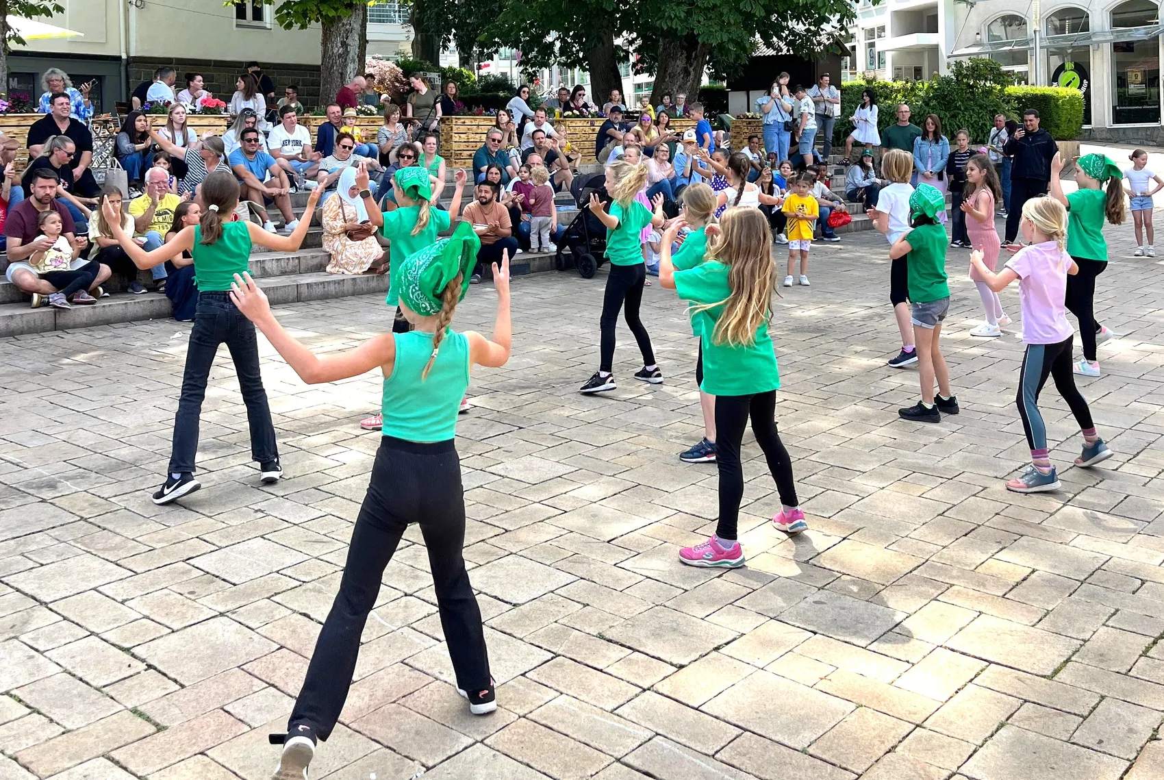 Tanzende Kinder und Jugendliche in grünen T-Shirts und schwarzen Hosen führen eine Choreografie auf einem gepflasterten Platz vor Zuschauern auf. Im Hintergrund sind Bäume und Gebäude zu sehen.