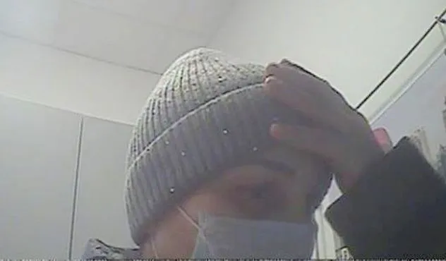Person mit Strickmütze und Mundschutz verdeckt das Gesicht mit der Hand vor einer Überwachungskamera.