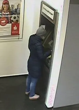 Eine Person mit grauer Kopfbedeckung und dunkler Jacke steht vor einem Geldautomaten und bedient ihn.