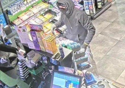 Maskierter Täter entwendet Zigarettenpackungen aus einem Regal an einer Kasse.