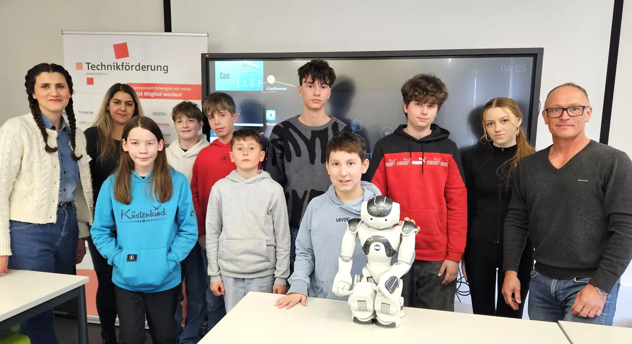 Gruppe von Kindern und Jugendlichen mit zwei Betreuern vor einem großen Bildschirm und einem humanoiden Roboter im Technikzentrum Lüdenscheid.
