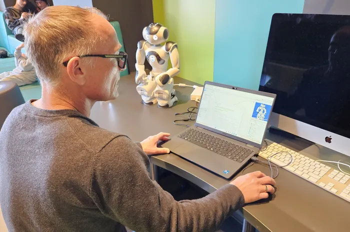 Ein Mann mit Brille sitzt vor einem Laptop und tippt, während im Hintergrund ein humanoider Roboter steht.