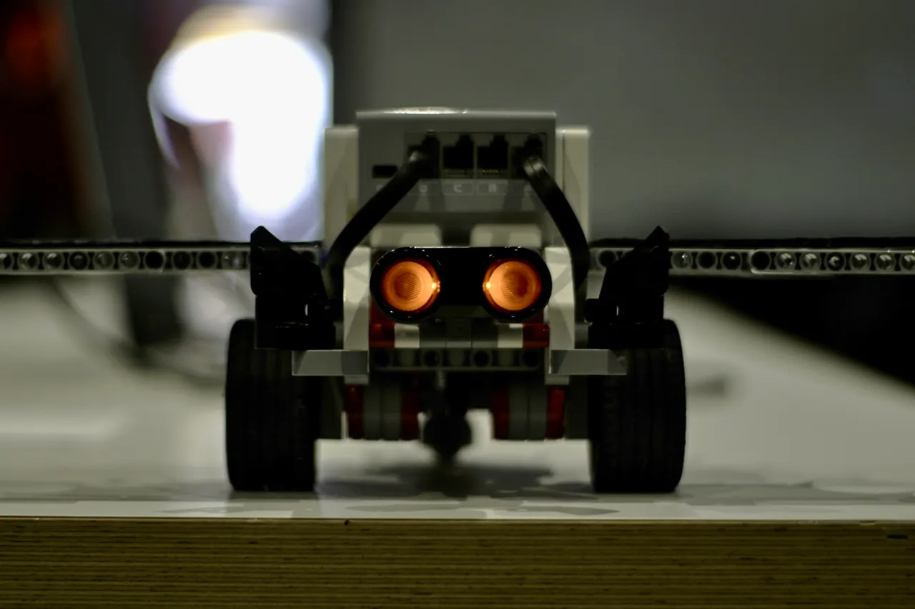Ein kleiner Roboter aus LEGO-Technik-Elementen mit zwei leuchtenden orangefarbenen Augen und schwarzen Rädern.