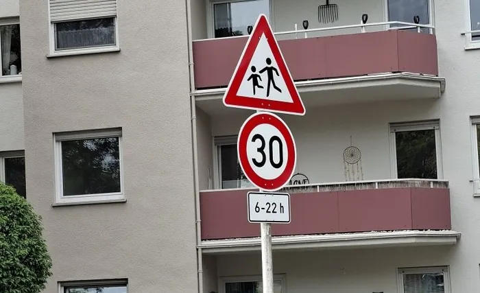 Verkehrsschilder mit einem Warnzeichen für Kinder, einer Geschwindigkeitsbegrenzung von 30 km/h und dem Zusatzschild „6–22 h“ vor einem Wohnhaus.