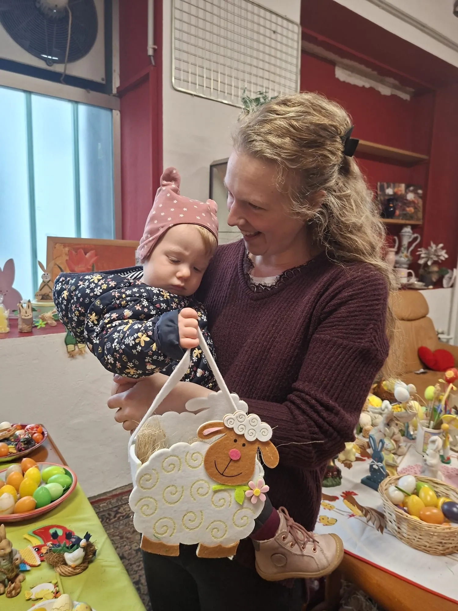 Eine Frau hält ein Baby auf dem Arm und präsentiert ein Osterkörbchen in Form eines Schafes. Im Hintergrund sind weitere Osterdekorationen zu sehen.