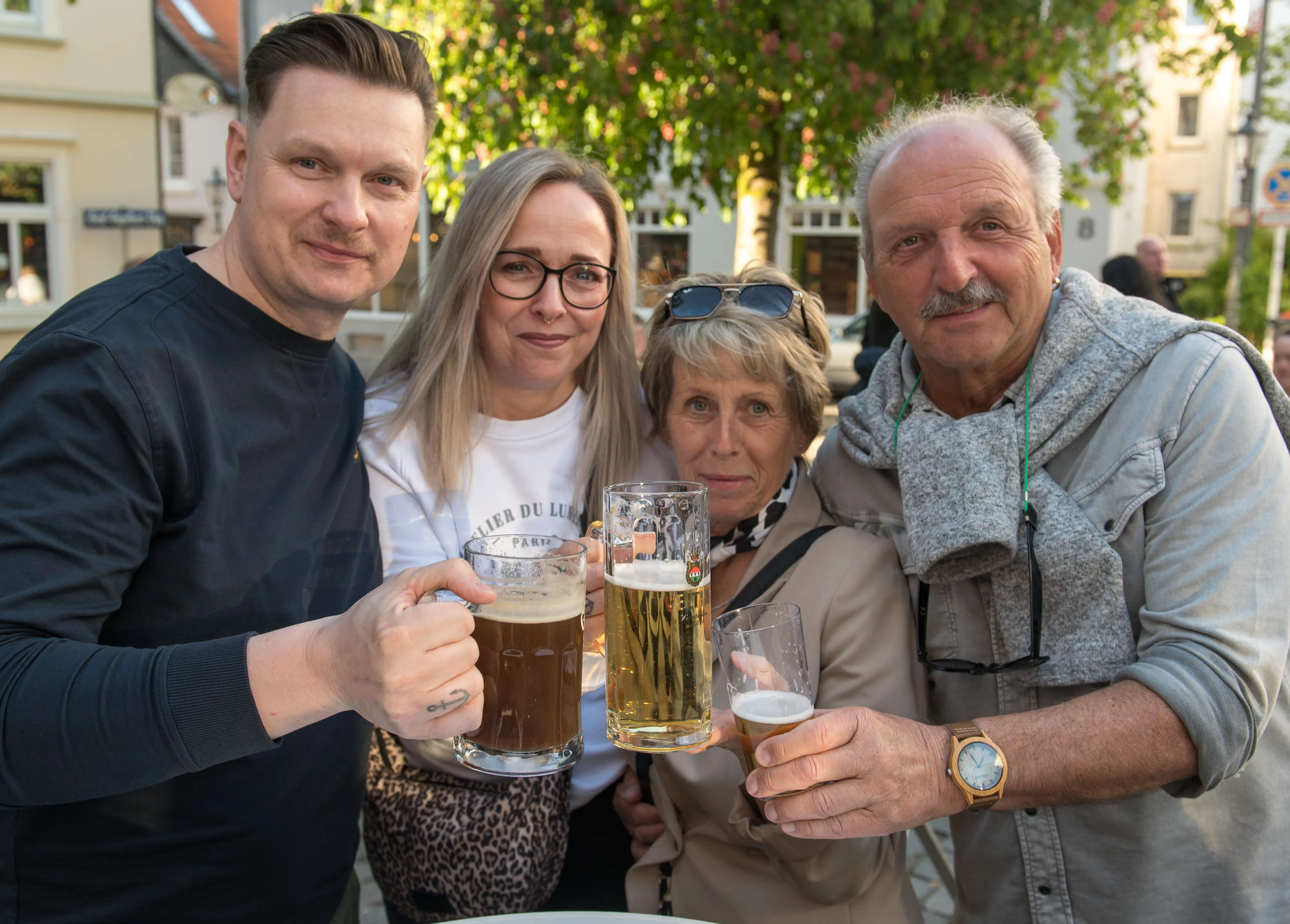 Vier Menschen stoßen mit Biergläsern an und lächeln in die Kamera. Im Hintergrund ist eine belebte Straße mit Geschäften und Gebäuden zu sehen.
