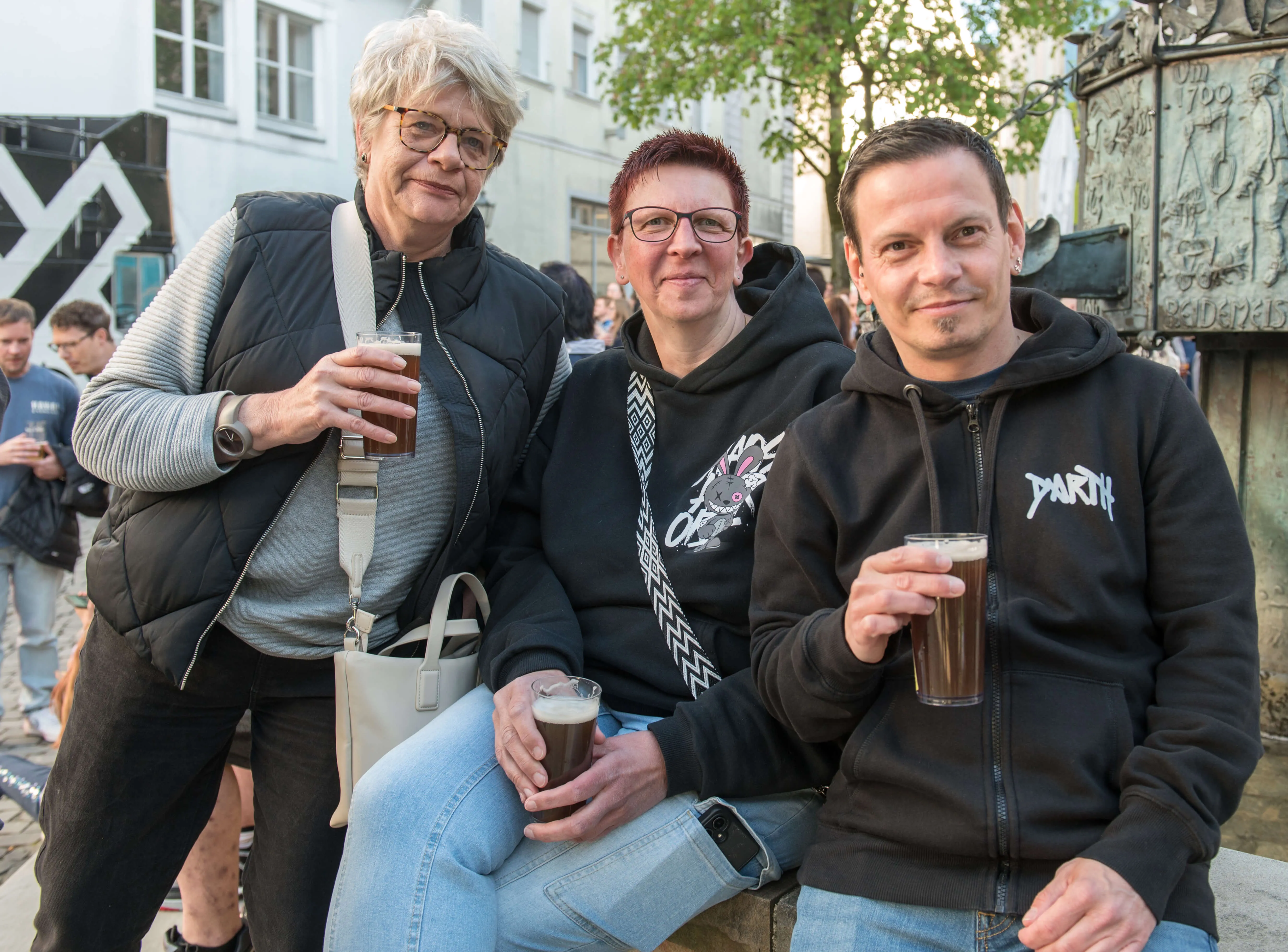 Drei lächelnde Menschen sitzen oder stehen eng zusammen und halten jeweils ein Glas Bier in der Hand. Im Hintergrund ist eine belebte Straße mit weiteren Menschen und Gebäuden zu sehen.