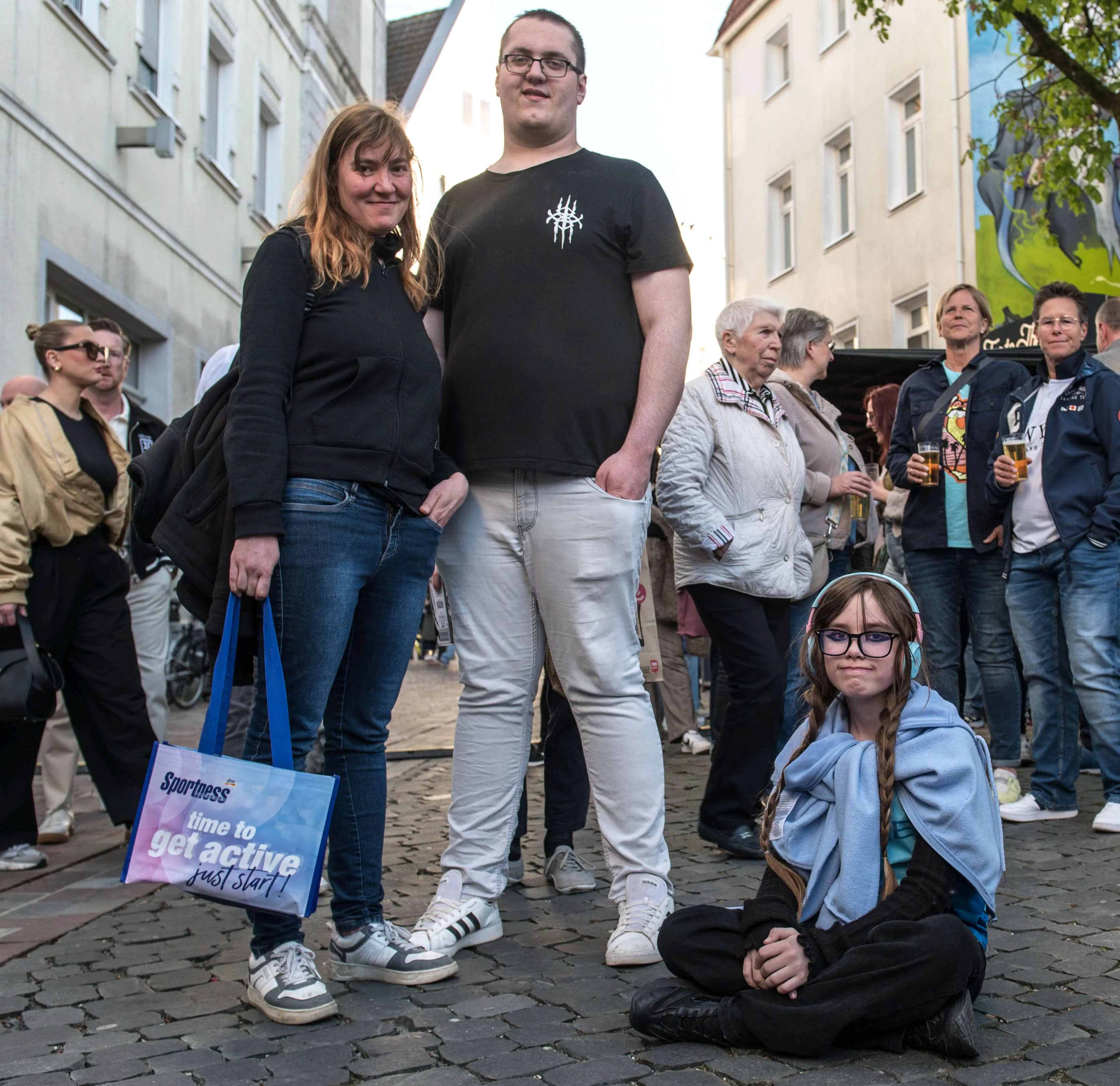 Zwei Personen stehen vor einer Gruppe von Menschen, während eine dritte Person mit Brille und blauer Jacke auf dem Kopfsteinpflaster sitzt. Im Hintergrund sind weitere Menschen und Geschäfte zu sehen.